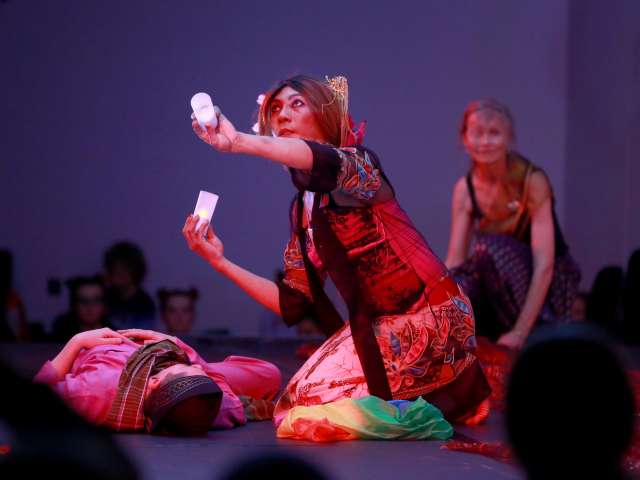 Three performers dressed in colourful Indian traditional costume demonstrate Bollywood style dance on a stage. A man lies on the floor on the left. A kneeling woman in the foreground moves her arms dramatically holding lights. A woman watches from the background. Silhouettes of the audience watch in foreground and background. 