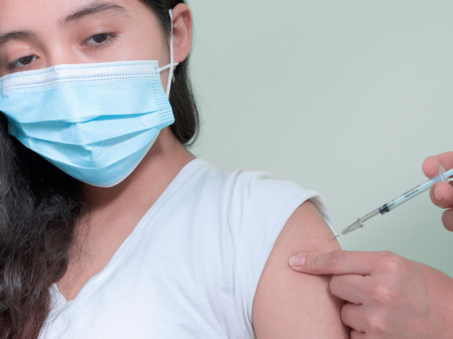 Teenage girl with dark long hair in pale v-necked top receives her vaccination in her left upper arm from a vaccinator on the right who is unseen apart from their hands.