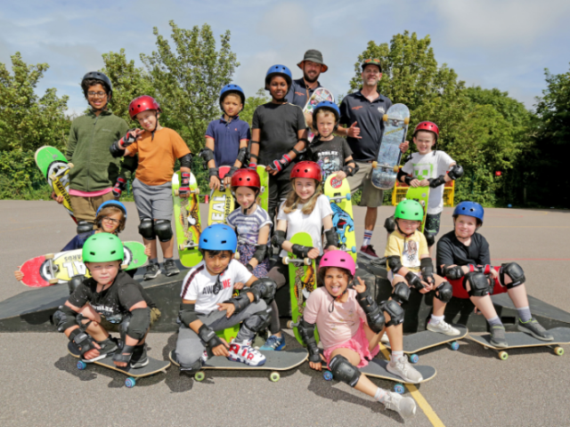 A large group of children, aged about 6 to 12, and their tutor pose with skateboards in sunny weather  