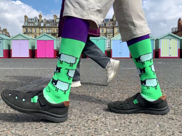 A man's legs walking in front of some beach huts. The man is wearing brightly coloured socks