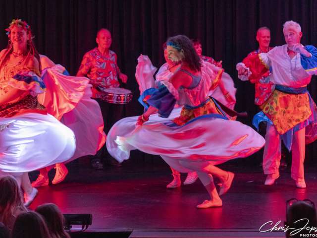 A group of women and men in flamboyant colourful costume perform on a lit stage. Two women in the foreground dance energetically, swirling their voluminous skirts. They are accompanied by male drummers in the background. 