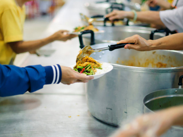 Hands holding up plates on left hand side with hands on right hand side dishing up food onto the plates from large metal pots
