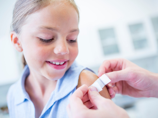 Young girl with long fair hair and a blue top getting a plaster put on her left arm