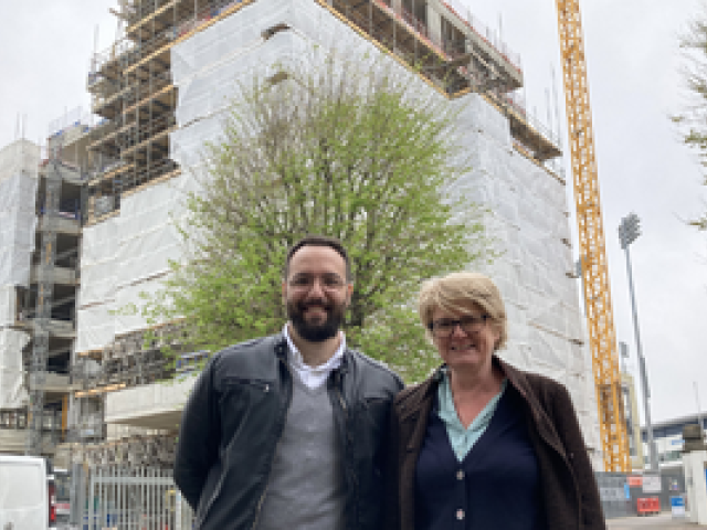 UCL student Ahmad Loukeh standing next to B&HCC Head of Planning Liz Hobden in front of a construction site for a tall block of flats. There is a tree immediately in front of the flats, which are clad in white material except for the top two floors. There is a yellow crane beside the flats and a floodlight pylon in the 1st Central County Cricket Ground in Hove behind that.
