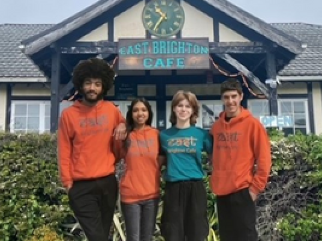 Two young men and two young women stand  in a line facing the viewer, three wearing orange tops. They stand in front of East Brighton Cafe which has a large clock in the peaked roof eaves above the cafe sign. A hedge runs in front of the cafe 