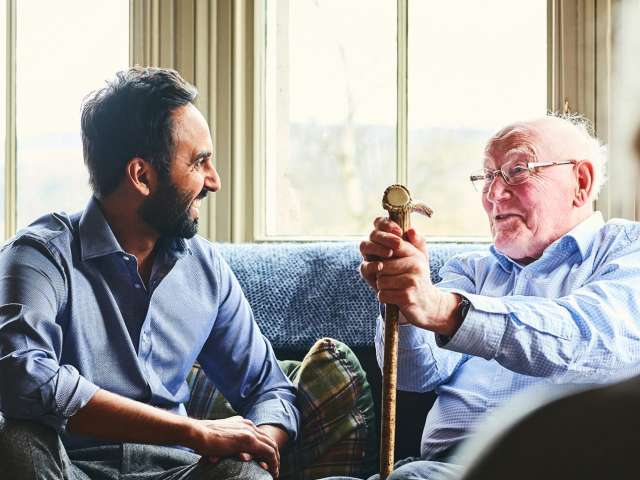 A  seated young man to the left speaks to an older seated man on the right who is resting his hands on a walking aid.