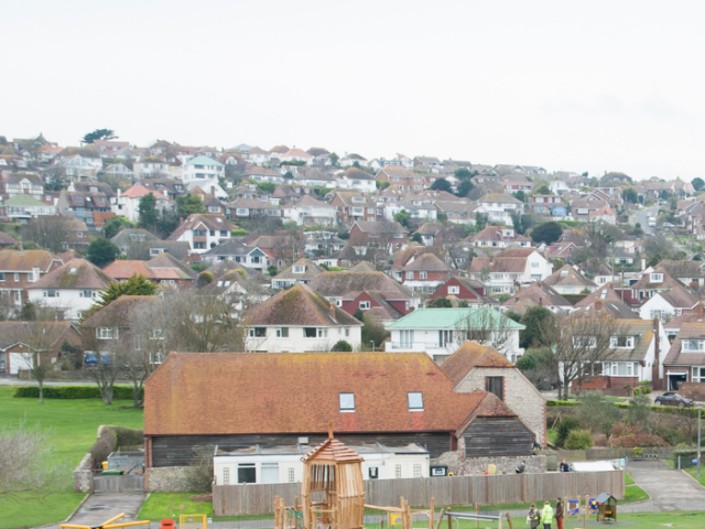 An aerial view of Saltdean with a childrens playground in the foreground, including a wooden climbing frame. Behind it there is a building in a park. Behind that houses rise up the hill to fill the horizon.