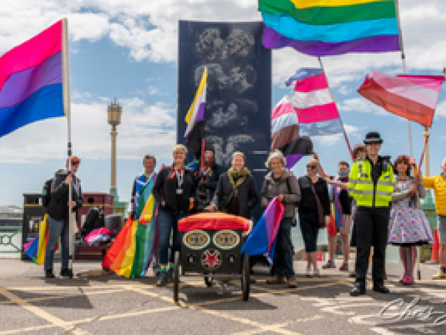 A group standing and waving LGBTQ+ flags in front of the kiss wall on Brighton seafront to mark IDAHOBiT in 2021. Photo courtesy of Chris Jepson.