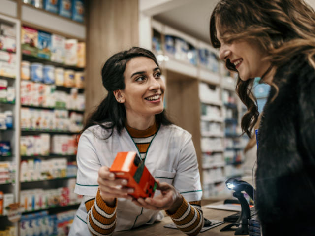 Female pharmacist white coat helps a female customer in a pharmacy by showing her a container of medication at the counter. Shelves of medication line the walls in the background   