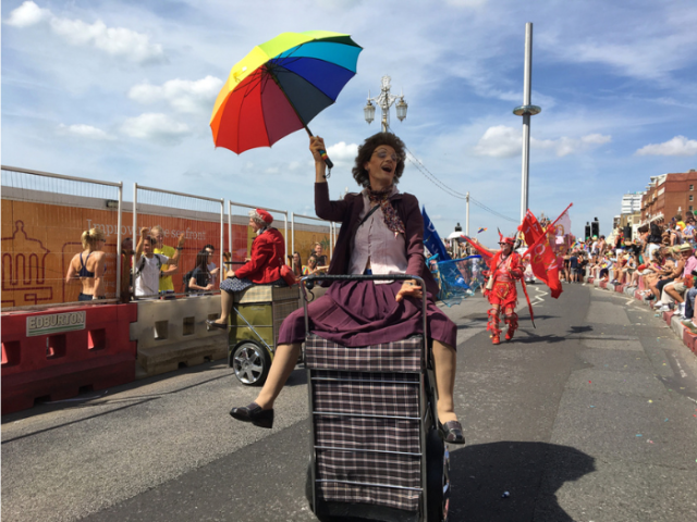A person sitting on a trolly holding a rainbow coloured umbrella in a parade