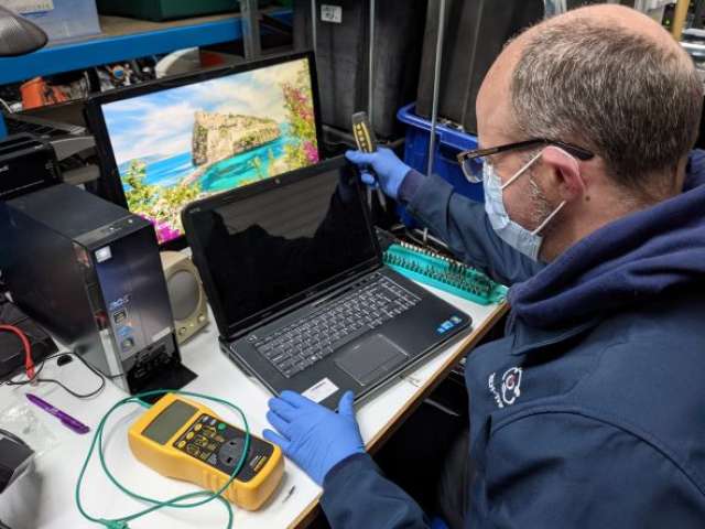 A man repairs a laptop, wearing a face mask and blue plastic gloves. He is holding a screwdriver. next to him is a yellow testing device.