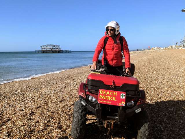A member of the Seafront team on patrol on Brighton Beach. He is riding a red quad bike with the words 'Beach Patrol' in red  on the front. He wears a red puffa jacket, white helmet and sunglasses. The sea and, in the distance, the West Pier, are behind him.