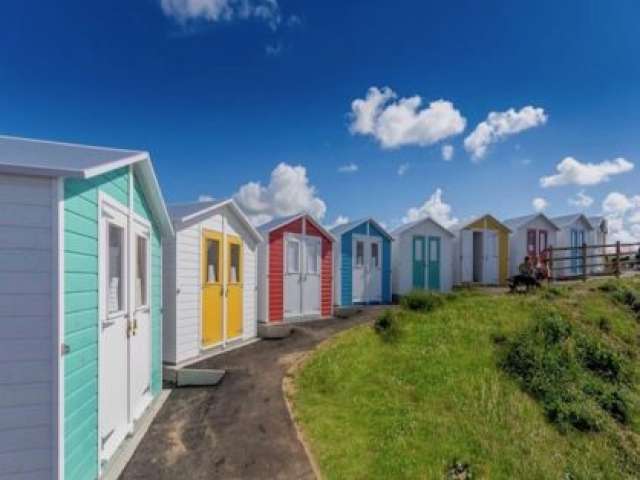 A row of white beach chalets with double doors. The doors or surrounding panels are painted in bright, primary colours.