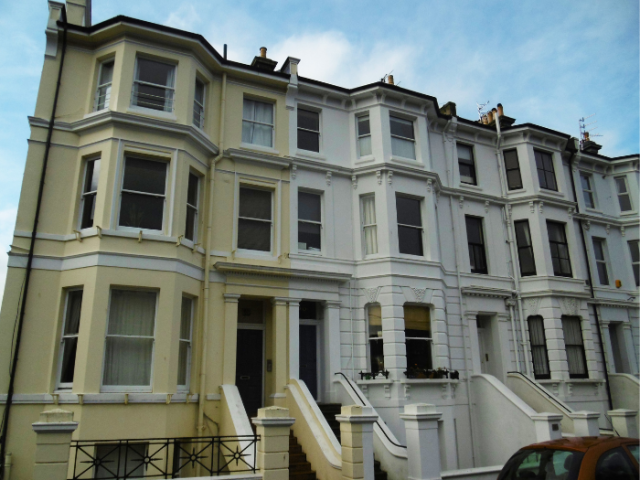 The picture shows a four-storey terrace of Victorian houses. The nearest is painted in an off-white colour, the rest in white. There are black rainwater downpipes and railings at street level. A red car is parked in the foreground. Steps lead up fom the street to the first floor front doors, with basements visible below.