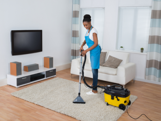 smiling woman using a carpet cleaner on a cream rug. The room she's in has a white sofa, TV on the wall and speakers on a white low height sideboard