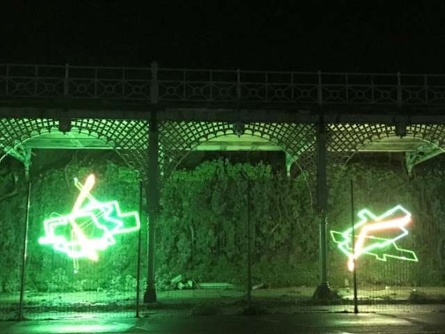 Under Madeira Terrace walk-way two arches are illuminated by multi-coloured neon installations of random shapes. The neon tubes light up the metal structure forming each arch and reflects on the wet street in front of them. Above the terrace the outline of the plants of the Green Wall can be seen in the darkness.
