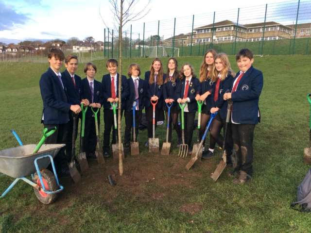 Year 8 students from Varndean School with spades and forks standing in a semi-circle having just planted one of four young trees on 'Stringer Way'