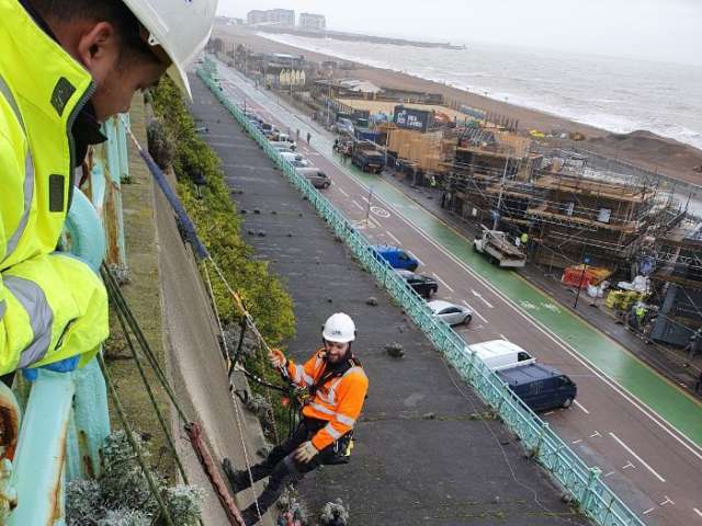 A climber dressed in an orange hi visability jacket, white helmet and black portective trousers is suspended by a rope and standing on the east cliff wall above Madeira Terrace. Below him you can see the Terrace platform, then the road below that with cars and vans parked in bays by the cliff wall. Further in the distance is the beach and scaffolding clad new buildings of the Sea Lanes development. A colleeague is wathcing the climber from above.