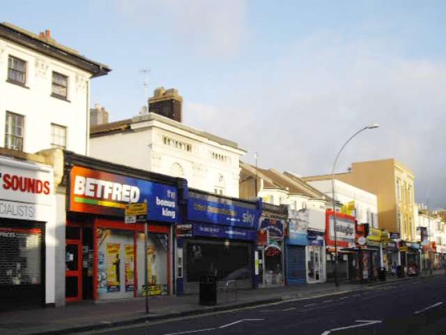 A row of shops on a busy high street streches away in to the distance as we look up the road. Above the shops are whtite painted first floors of houses. The road in front of the shops is empty of cars