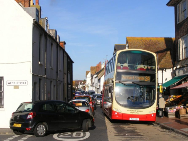 A bus is making its way down a narrow street lined with shops. As it passes a shop with a teal awning a black car is about to turn left into the opposite site of the road but uis waiting for the bus to pass