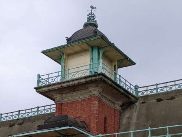 The ornate Victorian lift opening on Marine Parade rises up above the grey concrete Madeira Terrace wall. Beneath the blue railings surrounding the lift entrace the red brick lift shaft leans into the Terrace wall.
