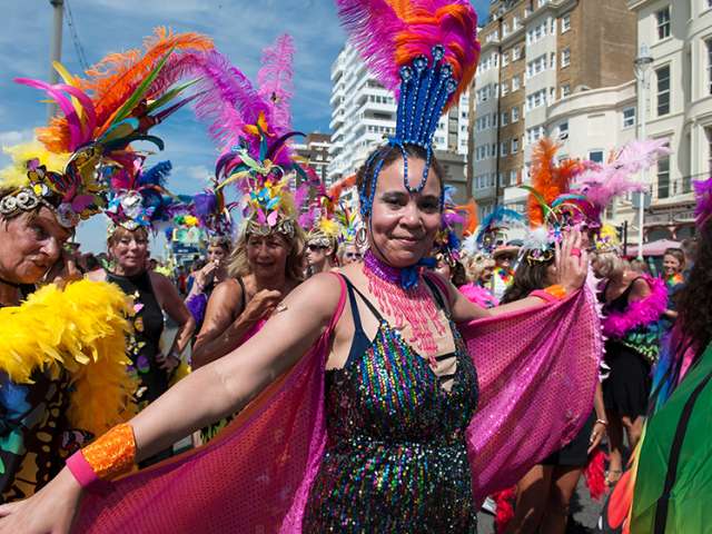People celebrating pride. A dancer is front and centre with a peacock plumage sequin top, a pink throw and a large feathered headpiece