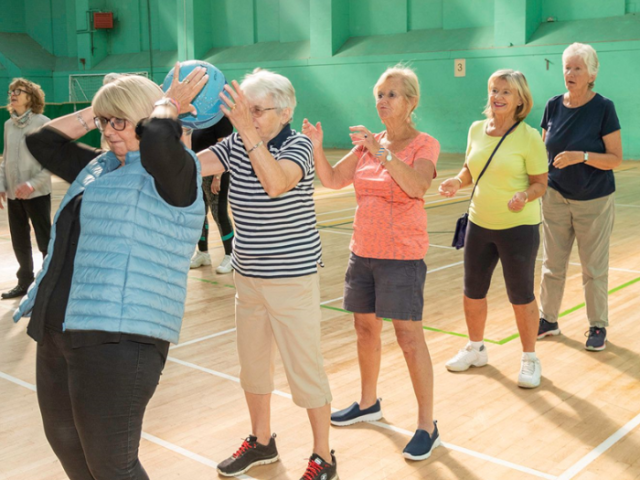 Photo of five older women in brightly coloured clothes standing in a line and passing a ball to each other over their heads