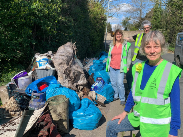 Photograph of 3 people wearing smiling at the camera next to a large pile of rubbish they have collected.