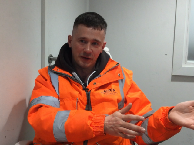 Photograph of young man with short hair looking at the camera. He is waving his arms and wearing a bright orange hi-vis jacket.