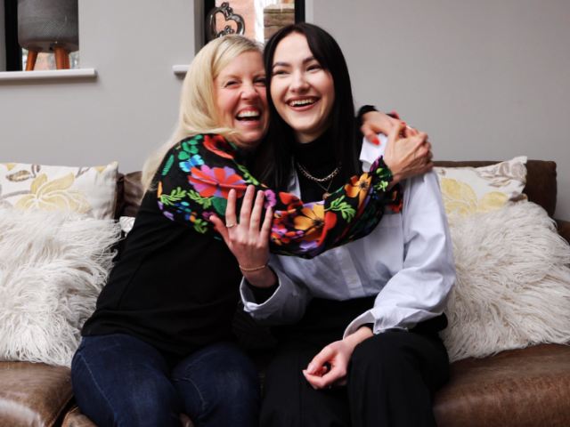 Image shows a photograph of a dark haired young girl being embraced by a blonde woman.