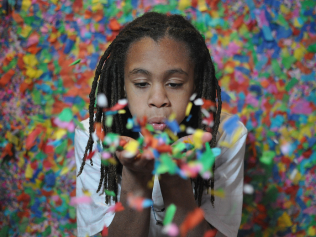 'A young black person with dreadlocks blows multi-coloured confetti towards the camera.'