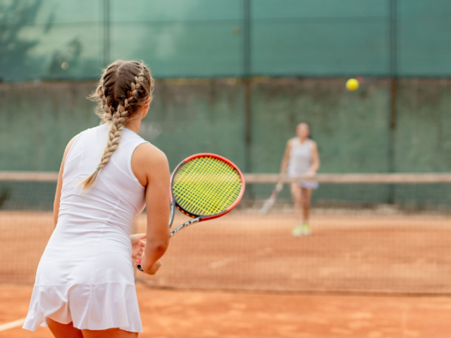 Photograph of a woman wearing white tennis clothes and holding a tennis racquet. Her opponent can be seen blurred in the background.