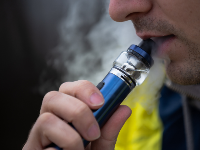 The image shows a lower portion of a man's face, exhaling smoke and holding a vape.
