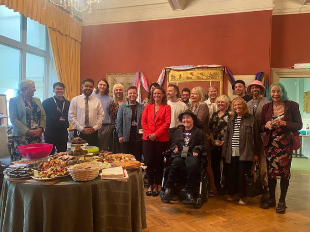 Image shows a group of people including Council Leader Bella Sankey in a red blazer, the Mayor of Brighton & Hove Jackie O'Quinn, a number of councillors and Trans Pride activists, smiling in a room decorated with trans and LGBTQ+ flags.