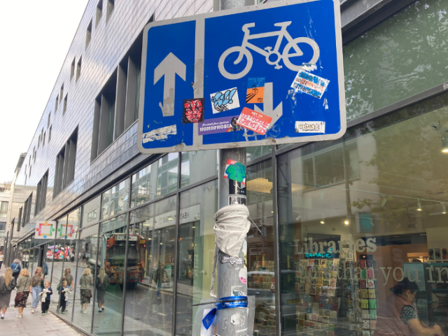 Photograph of a blue road sign with a bicycle on it. The sign is covered in stickers.