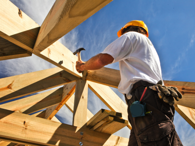 The image shows a construction worker wearing a hard hat and a tool belt, using a hammer on a roof beam.