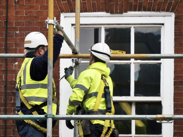 Two builders wearing white hard hats and yellow high visibility jackets are erecting scaffolding on the top floor of a house. The grey scaffolding poles have faded yellow paint markings and a white double glazed window can be seen behind the scaffolding.