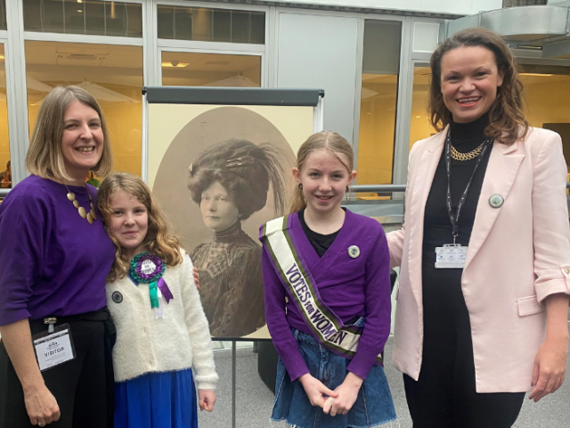 The image shows the Leader of the council, Councillor Bella Sankey with Mary Clarke's great great niece, Briony Goulden, and her two young daughters - Sylvie and Lena. Sylvie and Lena are wearing sashes and badges saying 'Votes for women'. Behind them is a picture of Mary Clarke.