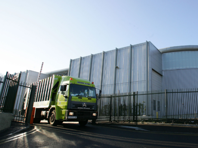 A yellow-fronted bin lorry leave a waste depot on a tarmac road that has black metal fencing on either side of the entrance. Double yellow lines cna be seen on the road and the top of a red and white painted barrier gate can be seen above thge top of the truck.Above the depot building behind the truck the sky is clear and an early morning sunshine can be seen.