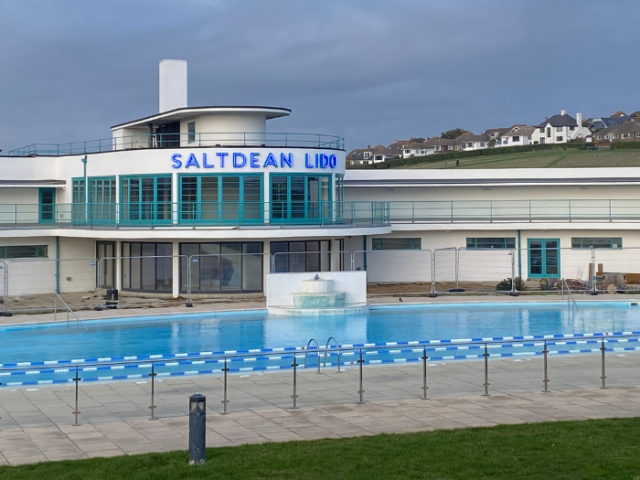The image shows Saltdean Lido building from distance on a cloudy day.