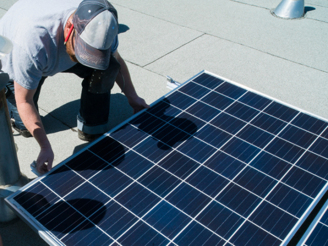 A man in a grey T shirt and jeans is moving a solar panel into positions on a light grey roof surface. He's wearing a grey and blac cap, the peak of which obsures his face as he looks down. He has a red pencil behind his right ear.