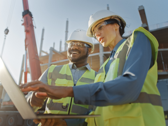 A man and a woman in hi-viz vests and hard hats on a building site checking a computer