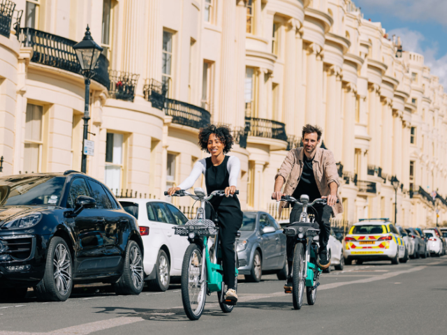 Two people riding bikes on a road with terraced houses and cars in the background"