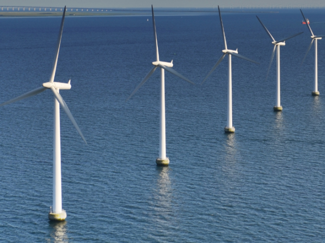 5 white 3-armed wind turbines stand in a line in the sea stretching away from right to left of the viewer.