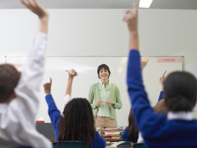 A school classroom from the back looking forward to a teacher stood at the front asking pupils questions. Three children in the foreground have their hands up to answer.
