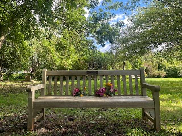 Wooden memorial bench with plaque in the middle. Flowers on seat of bench. In front of a large green lawn and green trees.