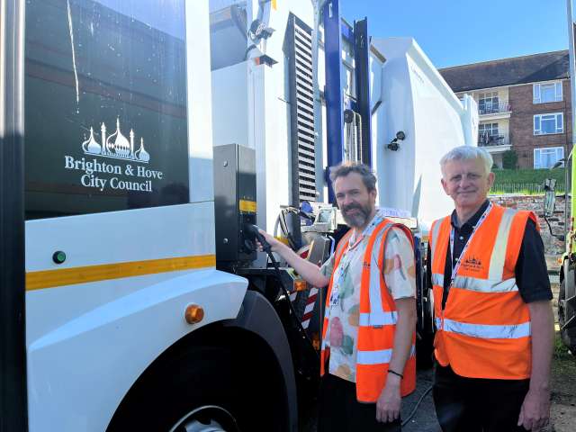 Councillor Tim Rowkins putting filling up the new electric truck for collecting waste for communal bins with councillor Trevor Muten standing next to him. Both are wearing orange high viz jackets