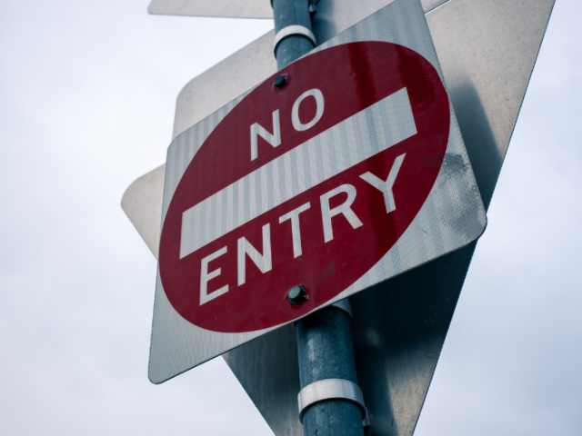 A road sign of a red circle with white bar horizontally across the middle and the words "No" and "Entry" above and below it. The sign is on a metal pole with a blue sky behind it. 