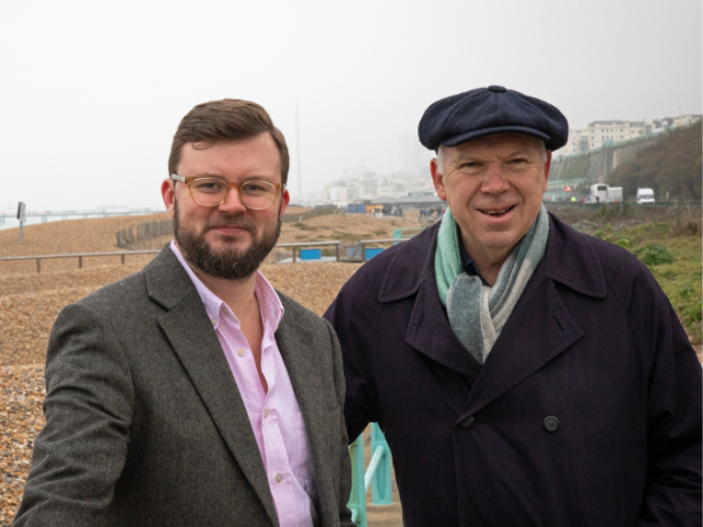 Two men are smiling and looking into the camera posing in front of a pebbled beach. One has a beard and glasses. The other is wearing a large cap and a blue scarf. Behind them the seafront of East Brighton can be seen stretching to the pier.