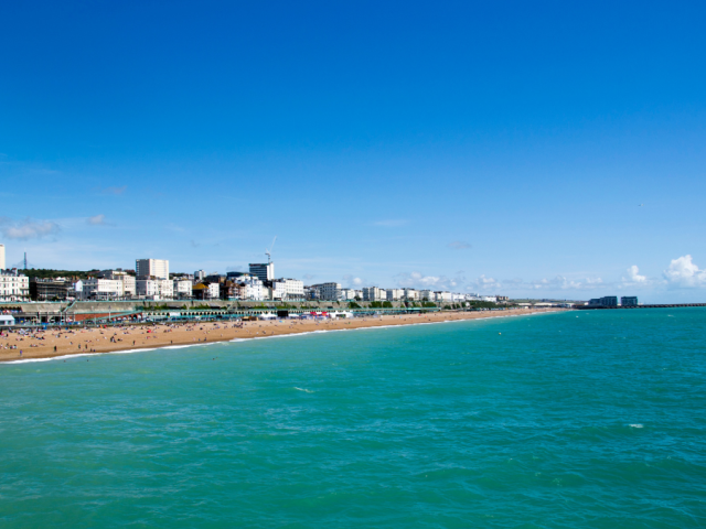 Brighton seafront with a blue sky and sea, with the beach on the left-hand side and buildings in the distance